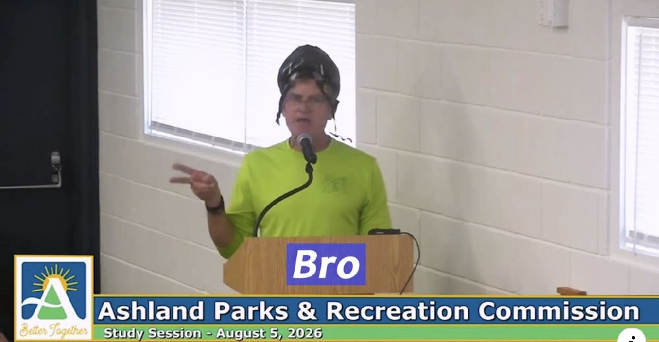 A man at a public-comment podium in a lime cycling jersey and a bike helmet worn over a wig, gesturing mid-sentence into the microphone. The meeting broadcast's lower third reads Ashland Parks & Recreation Commission, Study Session, August 5, 2026, and a name plate over the podium reads simply: Bro.